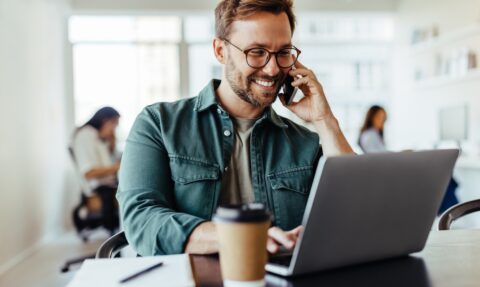 Male software designer speaking to a client on the phone while using a laptop. Happy young business man working in a co-working office.
